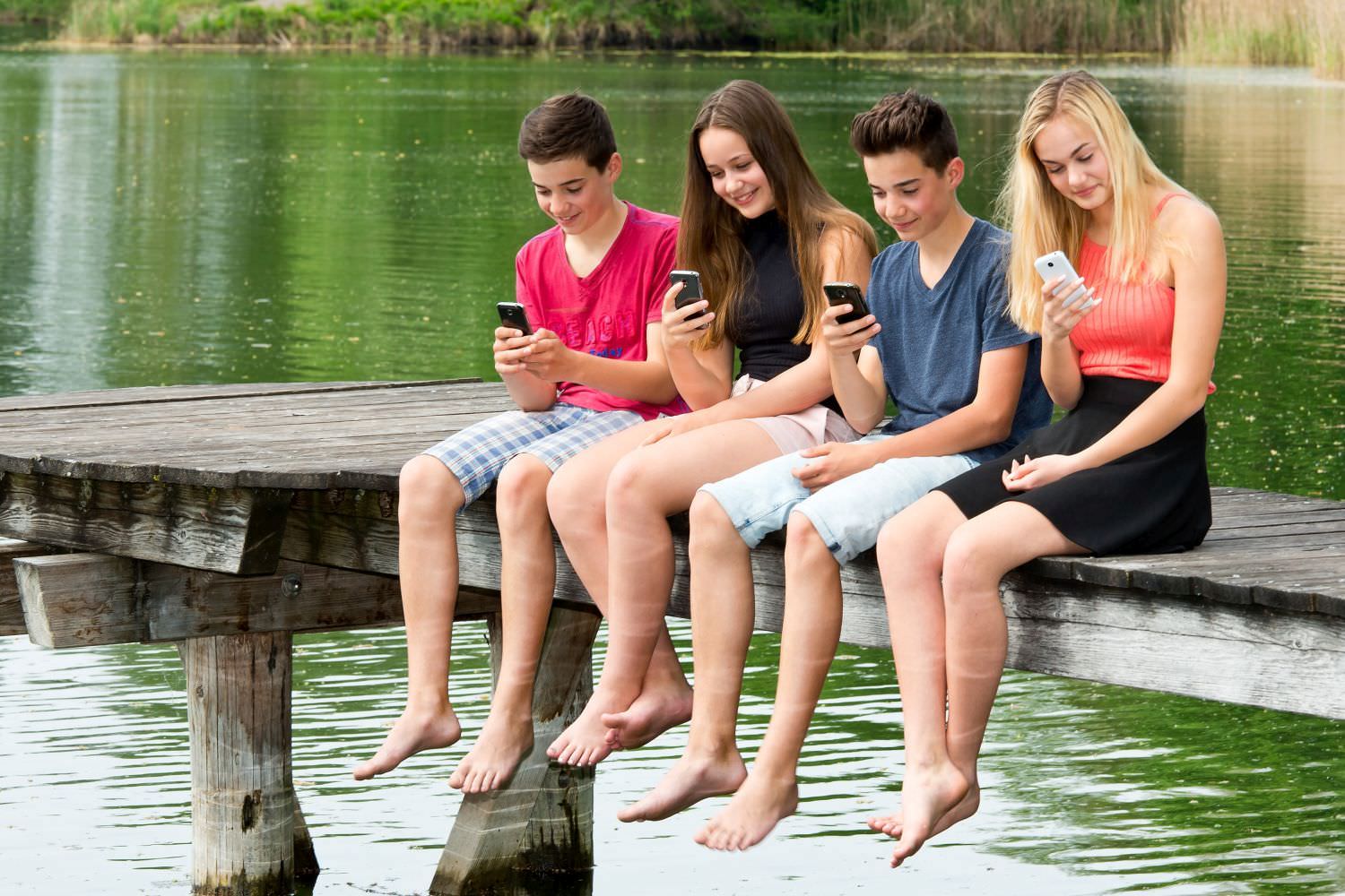 Bild zeigt: Vier Jugendliche sitzen auf einem Steg und schauen in ihre Smartphones