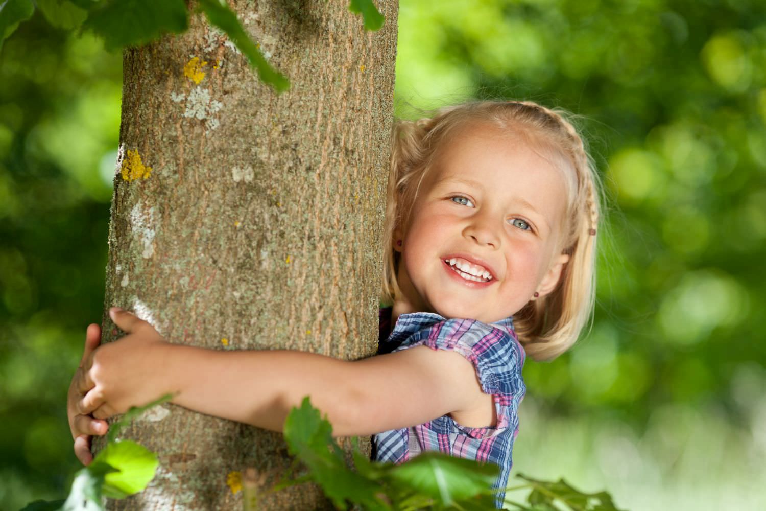 Bild zeigt: Ein kleines fröhliches Mädchen umarmt einen Baumstamm