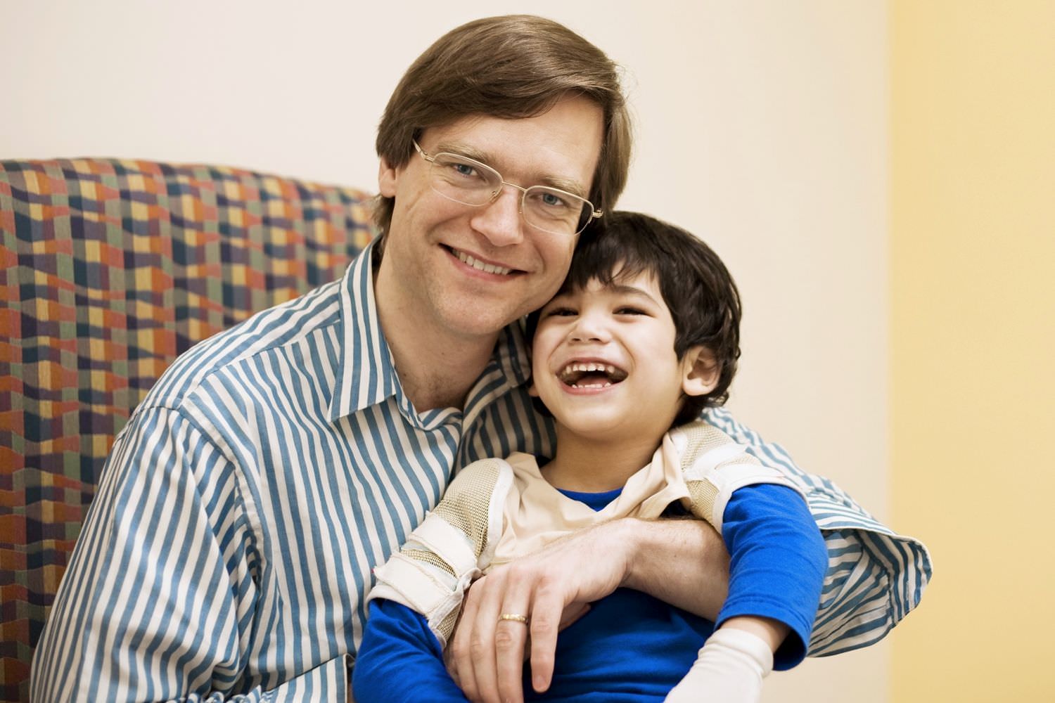 Bild zeigt: Junger glücklicher Vater sitzt mit seinem kleinen Sohn im Arm in einem Sessel