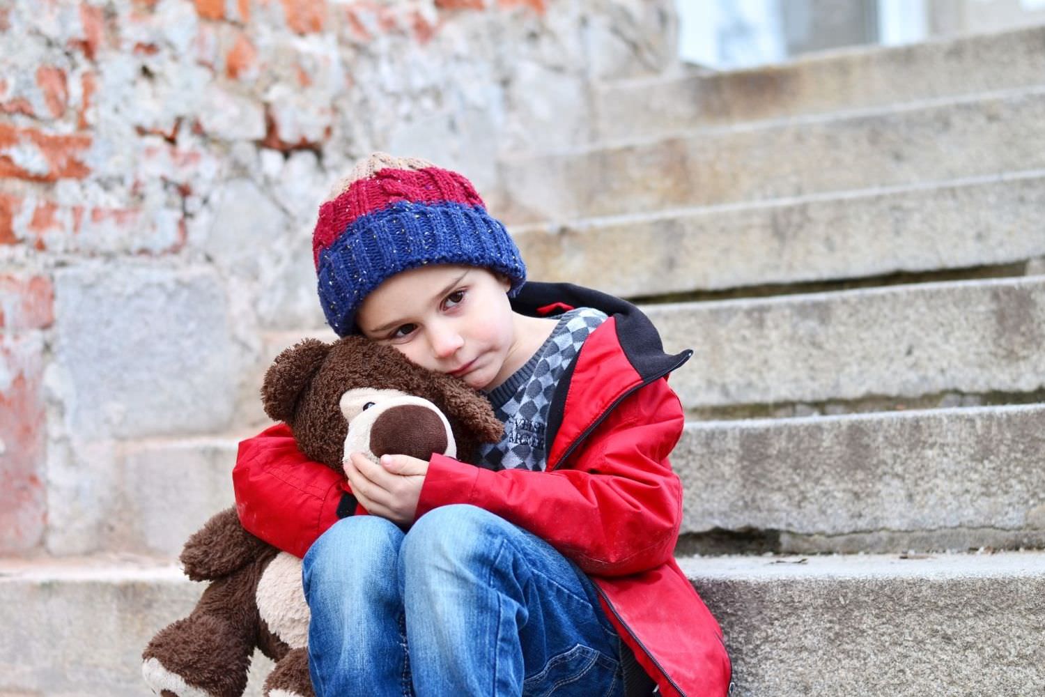 Bild zeigt: Kleiner trauriger Junge sitzt auf einer Steintreppe und hält sein Stofftier