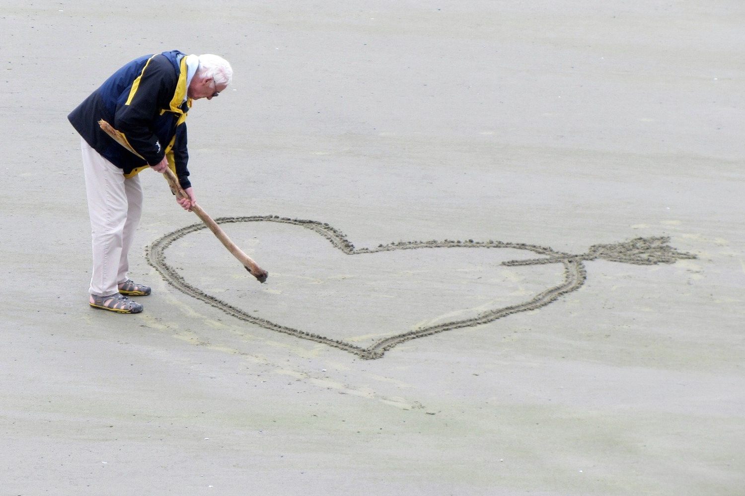 Bild zeigt: Alter Mann zeichnet mit Holzstock ein Herz in den Sand am Strand