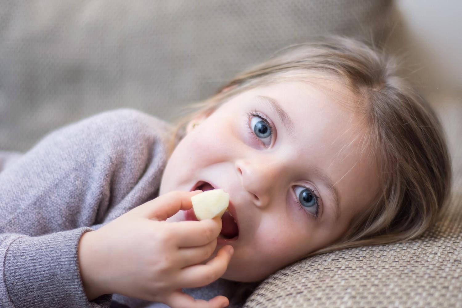 Bild zeigt: Kleines Mädchen liegt auf der Couch und isst ein Apfelstück