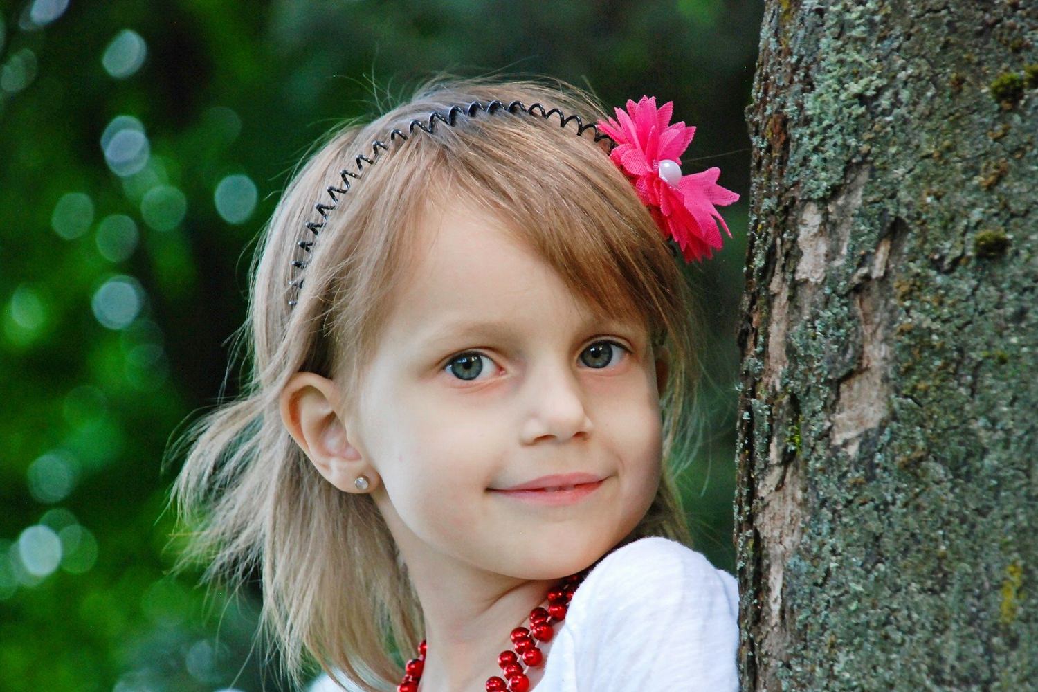Bild zeigt: Ein kleines Mädchen mit Blume im Haar lehnt an einen Baumstamm