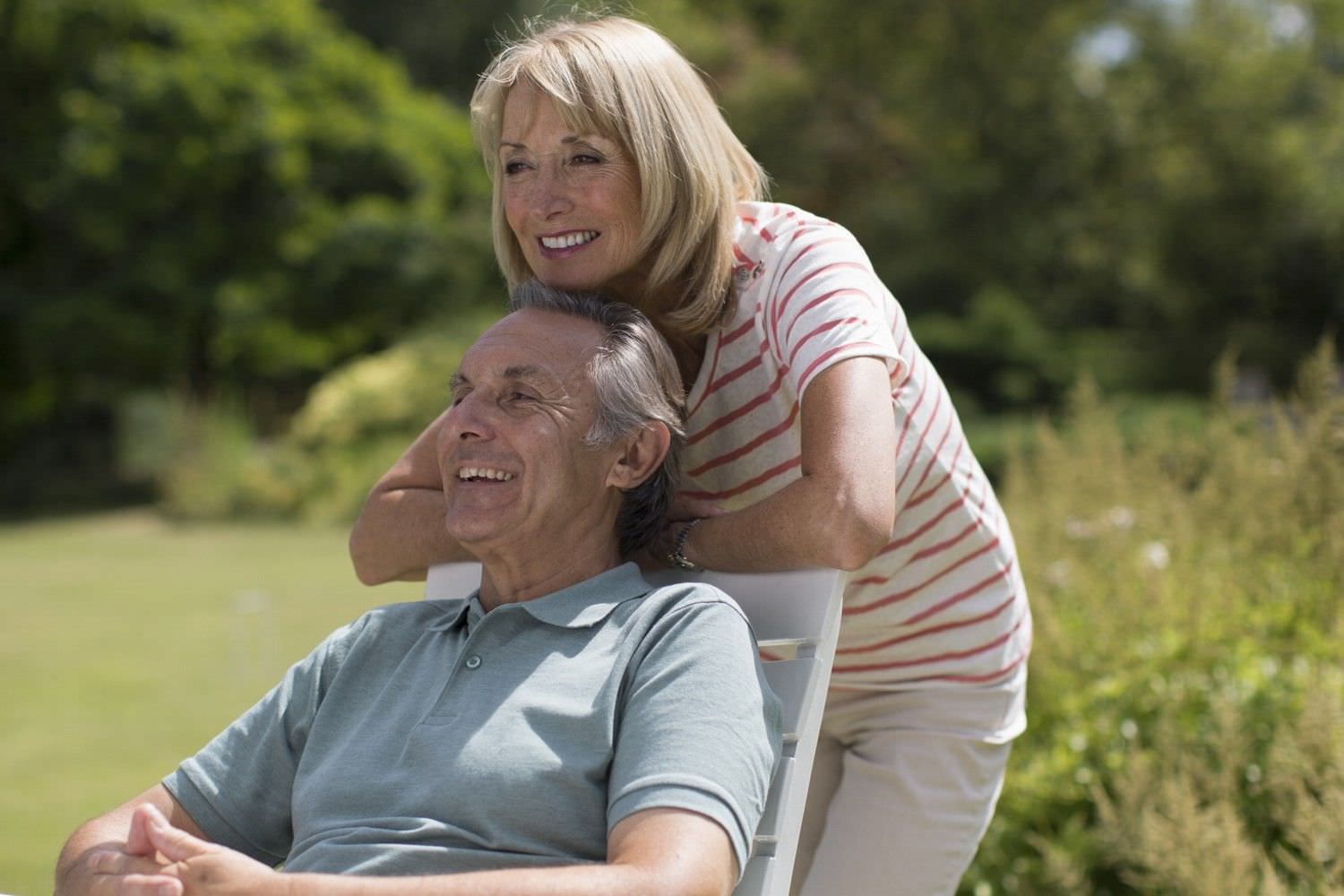 Bild zeigt: Ältere glückliche Frau steht draußen hinter dem Stuhl ihres Mannes