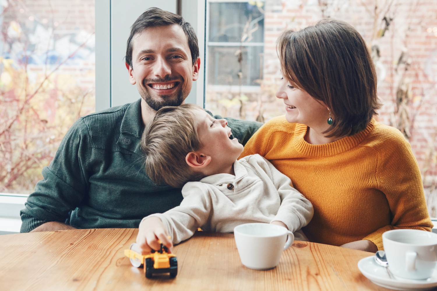 Bild zeigt: Vater, Mutter und Kleinkind sitzen fröhlich an einem Tisch mit Kaffeetassen